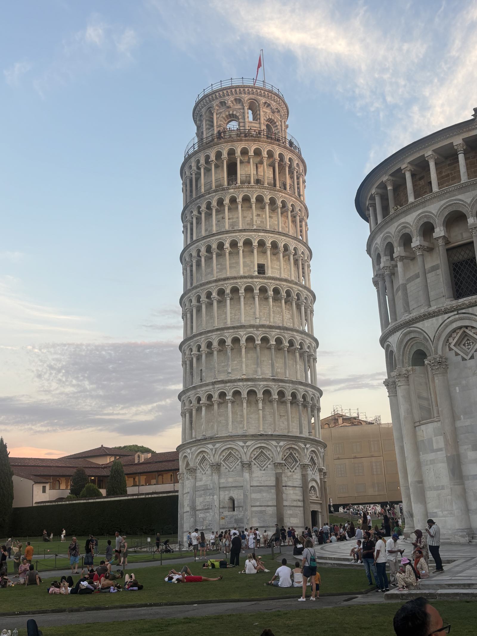 Der schiefe Turm von Pisa und Teile des Baptisteriums im Abendlicht, mit Besuchern auf der Wiese.