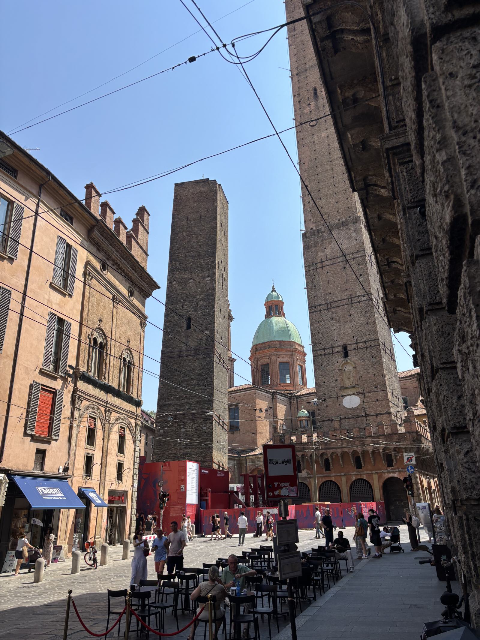 Zwei schiefe, hohe Backsteintürme in Bologna mit einer Kirche im Hintergrund und belebter Straße davor mit Cafétischen.