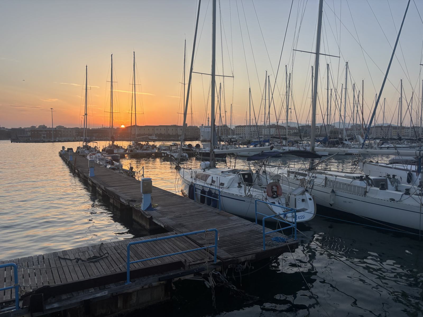 Sonnenaufgang über dem Hafen von Livorno mit an einem Holzsteg liegenden Segelbooten und der Stadtkulisse im Hintergrund. Das Wasser ist ruhig und spiegelt die Farben des Himmels wider.