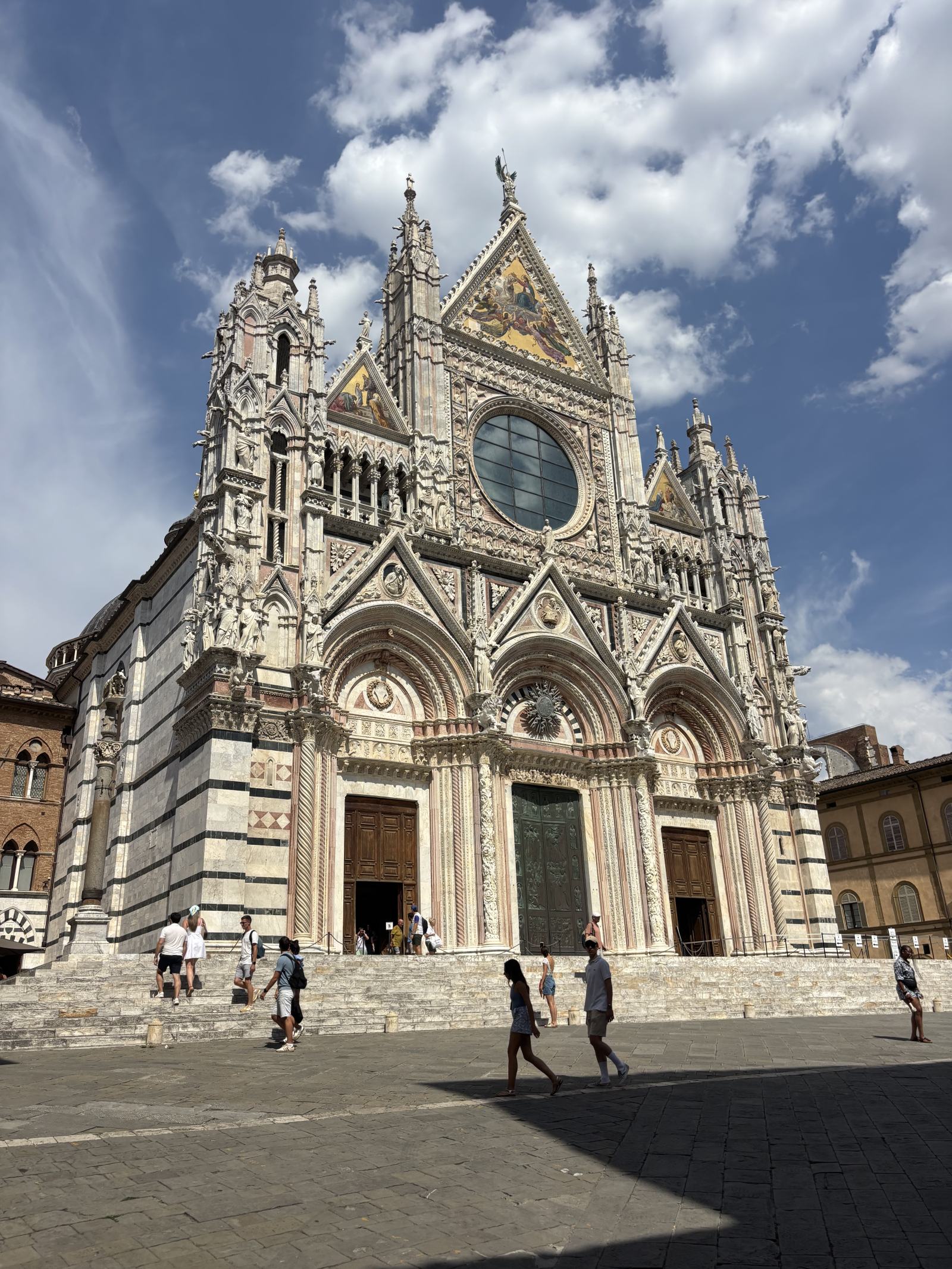 Die historische Fassade des Doms von Siena mit seiner markanten schwarz-weißen Marmorverkleidung, goldenen Mosaiken und einem großen runden Fenster. Mehrere Personen sind auf dem Vorplatz und den Stufen zu sehen.